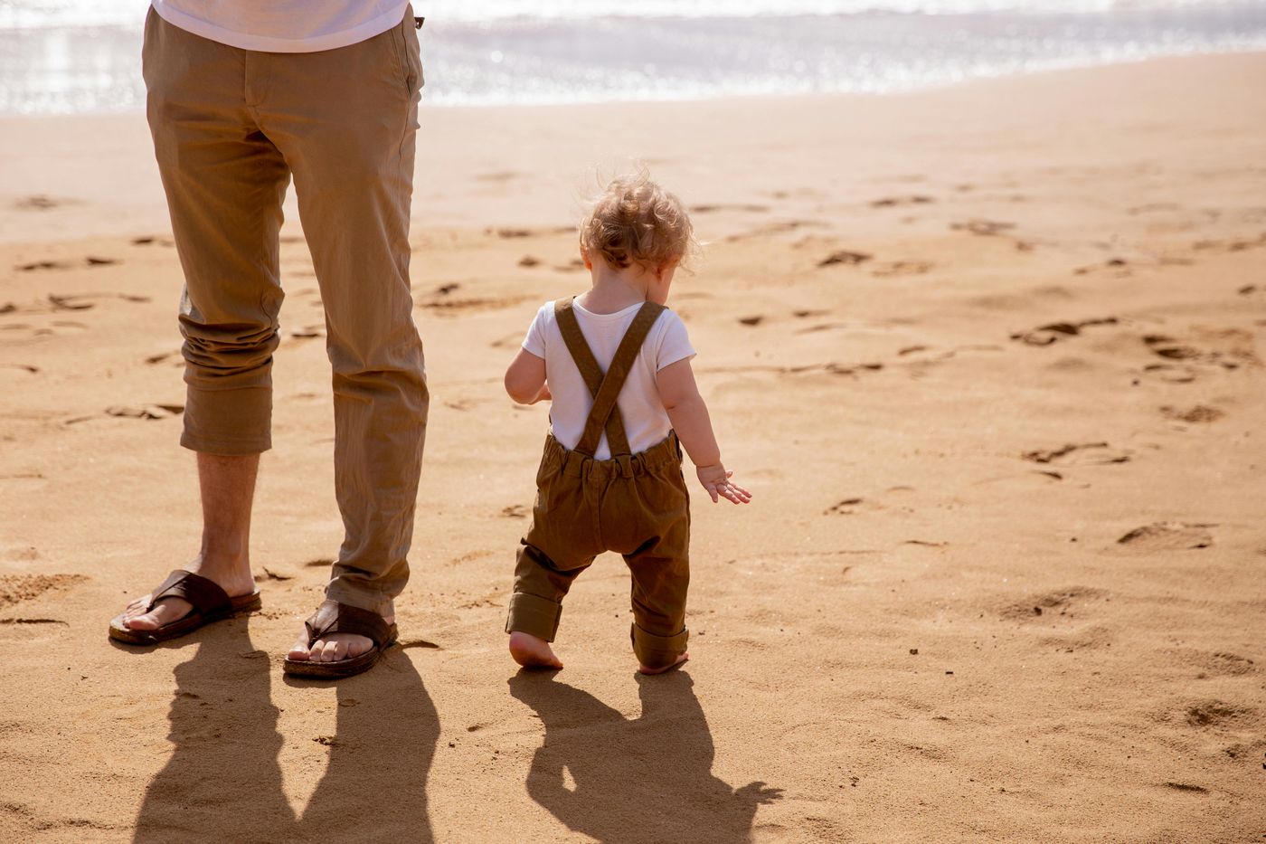 Kleinkind läuft neben seinem Papa barfuß über den Strand